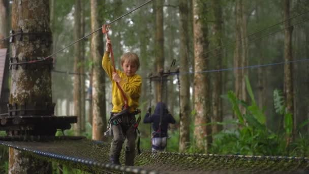 Superslowmotion shot van een jongetje in een veiligheidsharnas klimt op een route in boomtoppen in een forest Adventure Park. Hij klimt op een hoog touwparcours. Openlucht amusementscentrum met klimactiviteiten — Stockvideo