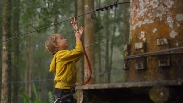 Superslowmotion skott av en liten pojke i en säkerhetssele klättrar på en rutt i trädtopparna i en skog äventyrspark. Han klättrar på hög repspår. Nöjescentrum utomhus med klättrings aktiviteter — Stockvideo