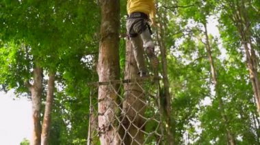 Bir güvenlik koşum küçük bir çocuk slowmotion çekim bir orman macera parkı ağaç tepelerinde bir zipline tırmanıyor. Zip hatları ve her türlü oluşan tırmanma faaliyetleri ile açık eğlence merkezi
