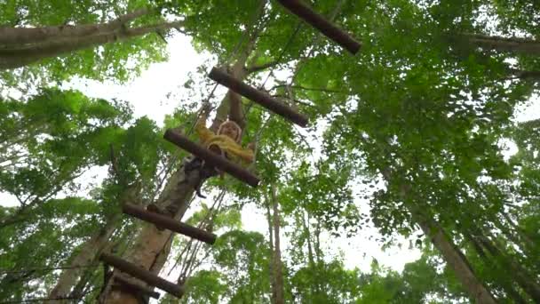 Повільний постріл маленького хлопчика в запобіжному в'язанні піднімається по маршруту на деревах в лісовому парку пригод. Він піднімається по високій мотузці. Відкритий центр розваг зі скелелазінням, що складається — стокове відео
