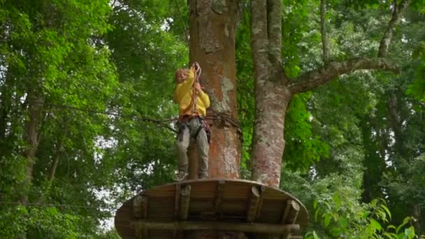 Slowmotion skott av en liten pojke i en säkerhetssele klättrar på en zipline i trädtopparna i en skog äventyrspark. Utomhus nöjescentrum med klättrings aktiviteter bestående av zip linjer och allehanda — Stockvideo