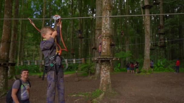 Superslowmotion skott av två små pojkar i en säkerhetssele klättra på en rutt i trädtopparna i en skog äventyrspark. De klättrar på hög repspår. Nöjescentrum utomhus med klättrings aktiviteter — Stockvideo