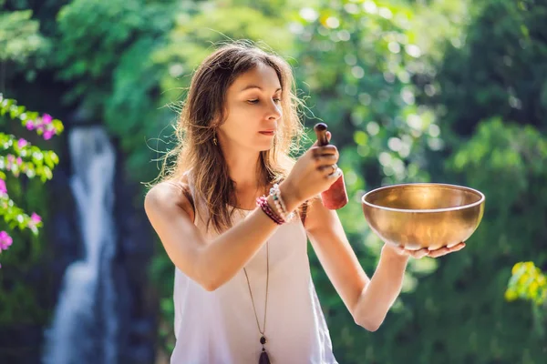 Mujer jugando en un cuenco tibetano mientras está sentada en una esterilla de yoga contra una cascada. Vintage tonificado. Hermosa chica con cuentas de mala meditación — Foto de Stock