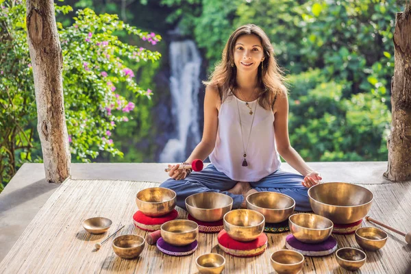 Donna che gioca sulla ciotola tibetana mentre siede sul tappetino yoga contro una cascata. Annata tonica. Bella ragazza con perline di mala meditando — Foto Stock