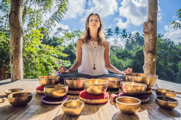 Frau spielt auf tibetischer Klangschale, während sie auf einer Yogamatte gegen einen Wasserfall sitzt. Jahrgangstonnen. schöne Mädchen mit Mala-Perlen meditieren — Stockfoto