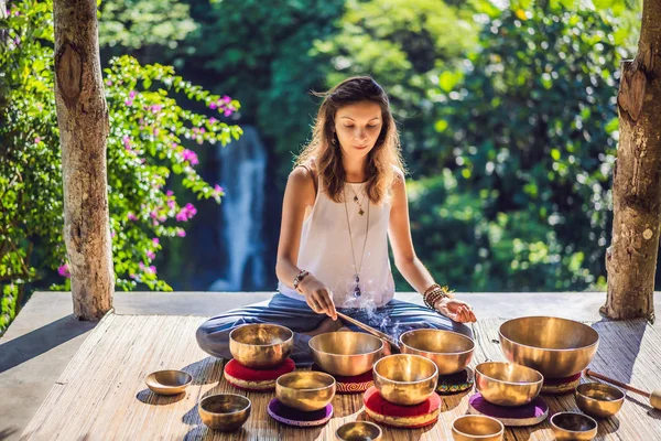 Donna che gioca sulla ciotola tibetana mentre siede sul tappetino yoga contro una cascata. Annata tonica. Bella ragazza con perline di mala meditando — Foto Stock