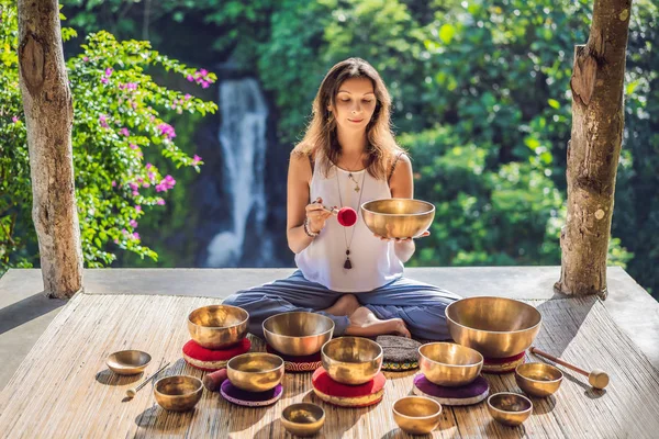 Frau spielt auf tibetischer Klangschale, während sie auf einer Yogamatte gegen einen Wasserfall sitzt. Jahrgangstonnen. schöne Mädchen mit Mala-Perlen meditieren — Stockfoto