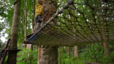 Bir güvenlik koşum küçük bir çocuk Superslowmotion çekim bir orman macera parkı ağaç tepelerinde bir rota üzerinde tırmanıyor. Yüksek halat patikalarına tırmanıyor. Tırmanma aktiviteleri ile açık hava eğlence merkezi