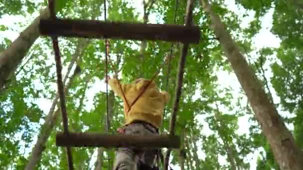 在森林探险公园的树梢上，一个穿着安全带的小男孩爬上了一条路线。他爬上高高的绳索。室外娱乐中心，登山活动由拉链和所有 — 图库视频影像