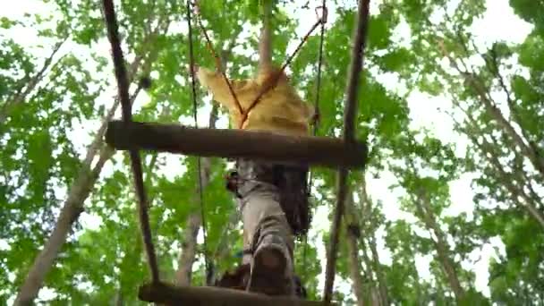 Un niño pequeño con un arnés de seguridad sube a una ruta en las copas de los árboles en un parque de aventura forestal. Se sube a un sendero de cuerda alta. Centro de diversiones al aire libre con actividades de escalada que consta de tirolesas y todo — Vídeos de Stock