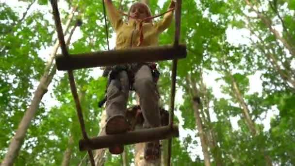 Menino em um arnês de segurança sobe em uma rota em copas de árvores em um parque de aventura florestal. Ele sobe em alta corda trilha. Centro de diversões ao ar livre com atividades de escalada consistindo de tirolesa e todos — Vídeo de Stock