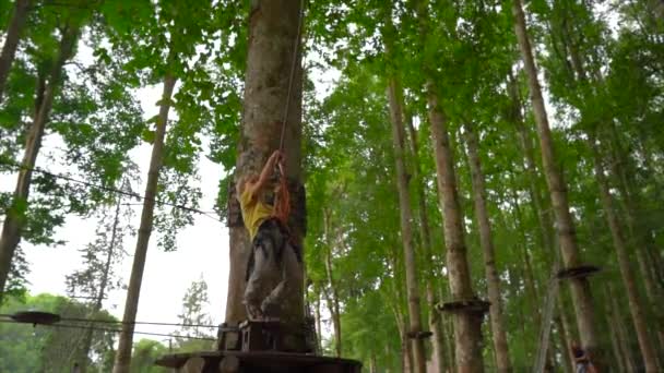在森林探险公园的树梢上，一个小男孩在安全带上的慢动作镜头。室外娱乐中心，包括拉链和各种攀岩活动 — 图库视频影像