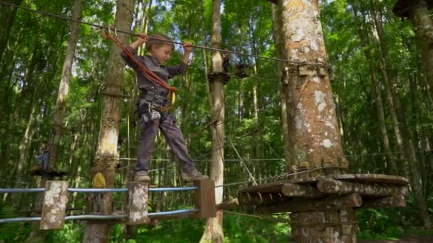 Filmagem em câmera lenta de um menino em um arnês de segurança sobe em uma rota em copas de árvores em um parque de aventura florestal. Ele sobe em alta corda trilha. Centro de diversões ao ar livre com atividades de escalada consistindo — Vídeo de Stock