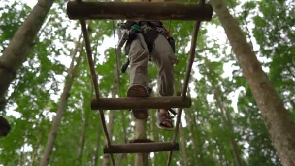 Superslowmotion skott av en liten pojke i en säkerhetssele klättrar på en rutt i trädtopparna i en skog äventyrspark. Han klättrar på hög repspår. Nöjescentrum utomhus med klättrings aktiviteter — Stockvideo