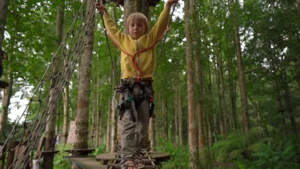 Superslowmotion scatto di un bambino con un'imbracatura di sicurezza sale su una via in cima ad un albero in un parco avventura foresta. Si arrampica su un sentiero ad alta corda. Centro di divertimento all'aperto con attività di arrampicata — Video Stock