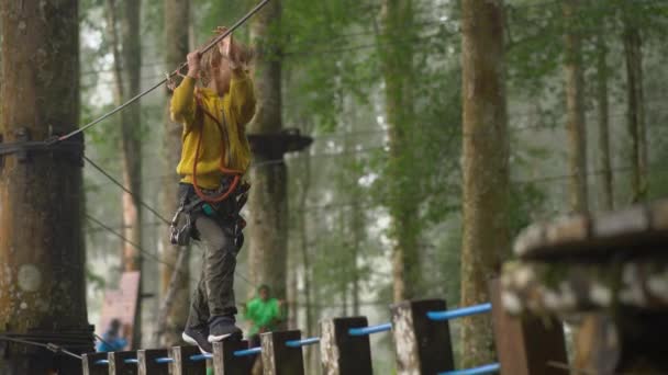 Liten pojke i en säkerhetssele klättrar på en rutt i trädtopparna i en skog äventyrspark. Han klättrar på hög repspår. Utomhus nöjescentrum med klättrings aktiviteter som består av zip linjer och alla — Stockvideo