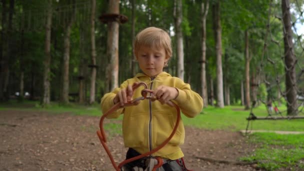 Το μικρό αγόρι σε ένα δασικό πάρκο περιπέτειας δείχνει την καλωδίωση ασφαλείας του. Υπαίθριο κέντρο ψυχαγωγίας με δραστηριότητες αναρρίχησης που αποτελούνται από γραμμές zip και όλα τα είδη εμποδίων για ενήλικες και παιδιά. Ενεργό — Αρχείο Βίντεο
