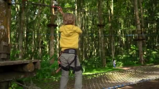 Menino em um arnês de segurança sobe em uma rota em um parque de aventura florestal. Ele sobe em alta corda trilha. Centro de diversões ao ar livre com atividades de escalada consistindo de tirolesa e todos os tipos de — Vídeo de Stock