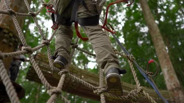 Liten pojke i en säkerhetssele klättrar på en rutt i trädtopparna i en skog äventyrspark. Han klättrar på hög repspår. Utomhus nöjescentrum med klättrings aktiviteter som består av zip linjer och alla — Stockvideo