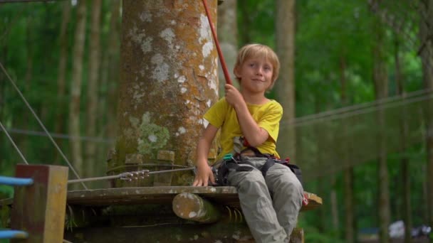 Superslowmotion skott av en liten pojke i en säkerhetssele på en rutt i en skog äventyrspark. Utomhus nöjescentrum med klättrings aktiviteter som består av zip linjer och alla typer av hinder för — Stockvideo