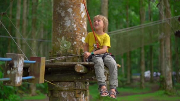 Superslowmotion shot van een jongetje in een veiligheidsharnas op een route in een forest Adventure Park. Openlucht amusementscentrum met klimactiviteiten bestaande uit zip-lijnen en allerlei obstakels voor — Stockvideo