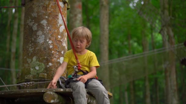 Superslowmotion skott av en liten pojke i en säkerhetssele på en rutt i en skog äventyrspark. Utomhus nöjescentrum med klättrings aktiviteter som består av zip linjer och alla typer av hinder för — Stockvideo