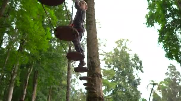 在森林探险公园的一条路线上，穿着安全带的小男孩爬上了一条路线。他爬上高高的绳索。室外娱乐中心，包括拉链和各种攀岩活动 — 图库视频影像