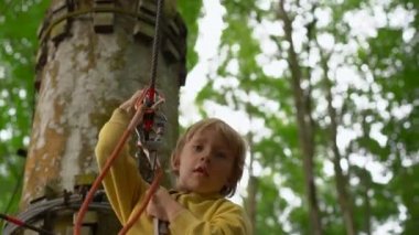 Bir güvenlik koşum küçük çocuk bir orman macera parkı ağaç tepelerinde bir zipline sürmek. Yüksek halat patikalarına tırmanıyor. Zip hatları ve tüm oluşan tırmanma faaliyetleri ile açık eğlence merkezi