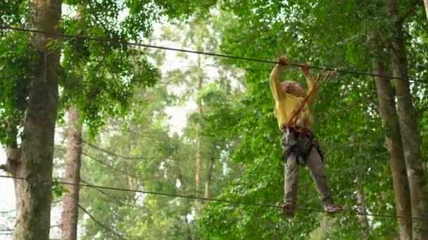 안전 벨트를 착용한 어린 소년이 숲 모험 공원의 나무 꼭대기에서 길을 따라 올라갑니다. 그는 높은 로프 트레일을 올라간다. 집라인과 모든 등산활동으로 구성된 야외 놀이센터 — 비디오