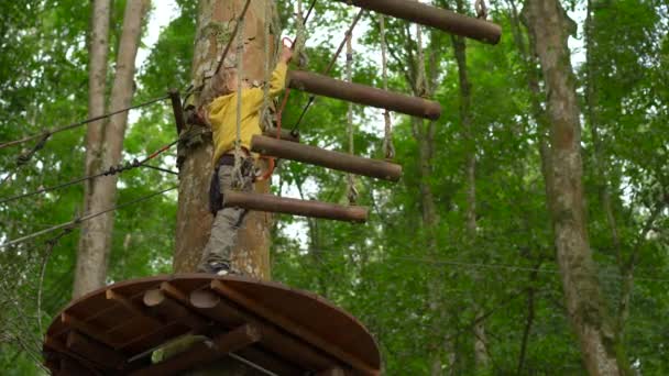 Маленький хлопчик у взутті піднімається на маршрут на деревах у лісовому парку пригод. Він піднімається по високій мотузці. Відкритий центр розваг зі скелелазінням, що складається з блискавок і всіх — стокове відео