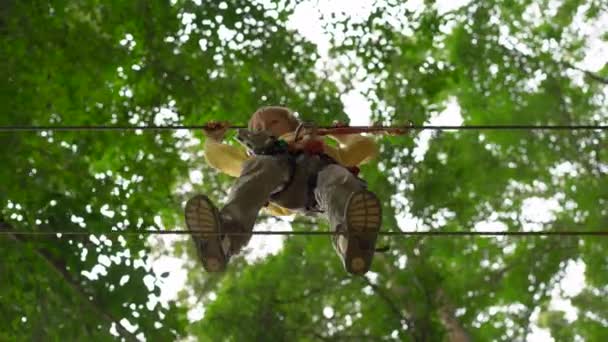 Liten pojke i en säkerhetssele klättrar på en rutt i en skog äventyrspark. Han klättrar på hög repspår. Utomhus nöjescentrum med klättrings aktiviteter bestående av zip linjer och alla typer av — Stockvideo