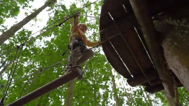 在森林探险公园的树梢上，一个小男孩在安全带上攀爬的慢动作镜头。他爬上高高的绳索。室外娱乐中心，包括登山活动 — 图库视频影像