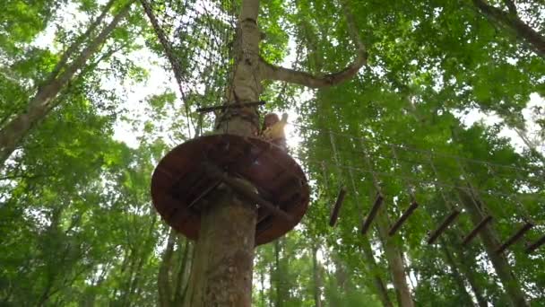 Slowmotion shot van een jongetje in een veiligheidsharnas klimt op een route in boomtoppen in een forest Adventure Park. Hij klimt op een hoog touwparcours. Openlucht amusementscentrum met klimactiviteiten bestaande uit — Stockvideo