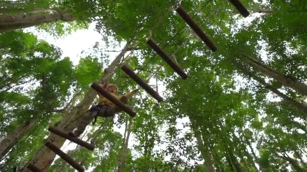 Slowmotion skott av en liten pojke i en säkerhetssele klättrar på en rutt i trädtopparna i en skog äventyrspark. Han klättrar på hög repspår. Nöjescentrum utomhus med klättrings aktiviteter bestående — Stockvideo