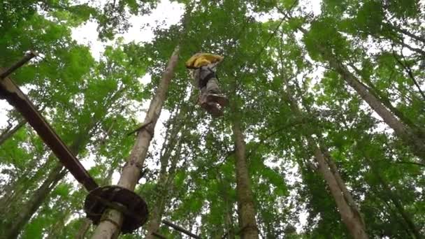 Filmagem em câmera lenta de um menino em um arnês de segurança sobe em uma rota em copas de árvores em um parque de aventura florestal. Ele sobe em alta corda trilha. Centro de diversões ao ar livre com atividades de escalada consistindo — Vídeo de Stock