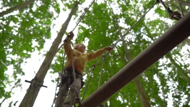 Superslowmotion disparo de un niño pequeño en un arnés de seguridad sube a una ruta en las copas de los árboles en un parque de aventura forestal. Se sube a un sendero de cuerda alta. Centro de diversiones al aire libre con actividades de escalada — Vídeo de stock