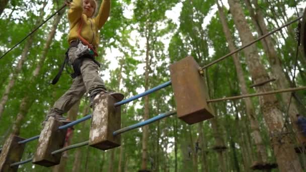 In Superzeitlupe klettert ein kleiner Junge im Sicherheitsgurt auf einer Route in Baumkronen eines Wald-Erlebnisparks. Er klettert auf einem Hochseilgarten. Outdoor-Vergnügungszentrum mit Kletteraktivitäten — Stockvideo