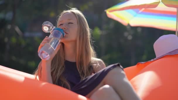 Mujer joven en una playa tropical se sienta en un sofá inflable y bebe agua de una botella de plástico de uso múltiple. Bebe más agua. Plástico reutilizable y reducir el concepto de residuos plásticos — Vídeos de Stock