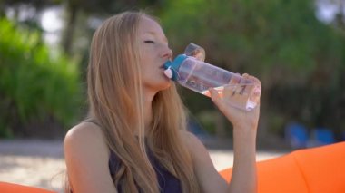Tropik bir plajda genç bir kadının Superslowmotion çekim şişme bir kanepe üzerinde oturur ve çok kullanılabilir plastik şişe su içecekler. Biraz daha su iç. Yeniden kullanılabilir plastik ve plastik atıkları azaltın