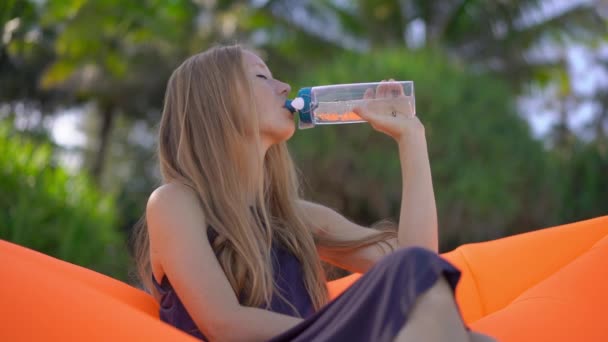 Суперслоументний знімок молодої жінки на тропічному пляжі сидить на надувному дивані і п'є воду з багаторазової пластикової пляшки. Пийте більше води. Багаторазові пластикові та пластмасові відходи — стокове відео