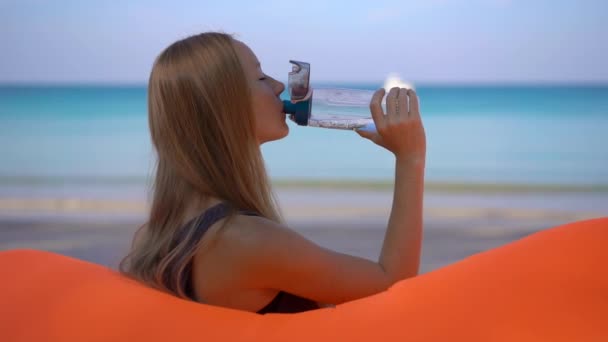 Superzeitlupenaufnahme einer jungen Frau an einem tropischen Strand, die auf einem aufblasbaren Sofa sitzt und Wasser aus einer mehrfach verwendbaren Plastikflasche trinkt. Mehr Wasser trinken. wiederverwendbarer Kunststoff und weniger Plastikmüll — Stockvideo