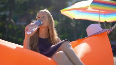 Tropik bir plajda genç bir kadının Superslowmotion çekim şişme bir kanepe üzerinde oturur ve çok kullanılabilir plastik şişe su içecekler. Biraz daha su iç. Yeniden kullanılabilir plastik ve plastik atıkları azaltın
