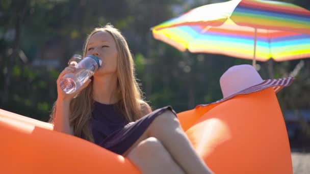 Superslowmotion disparo de una mujer joven en una playa tropical se sienta en un sofá inflable y bebe agua de una botella de plástico de uso múltiple. Bebe más agua. Plástico reutilizable y reducir los residuos plásticos — Vídeos de Stock