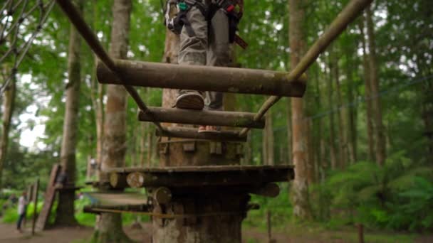 Superslowmotion shot of a little boy in a safety harness climbs on a route in treetops in a forest adventure park. He climbs on high rope trail. Outdoor amusement center with climbing activities — Stock Video