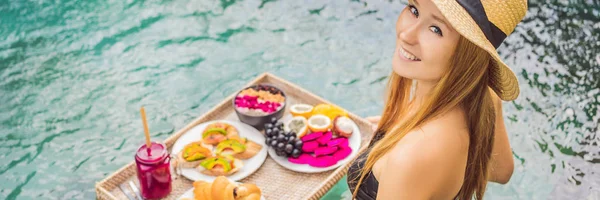 BANNER, LONG FORMAT Breakfast tray in swimming pool, floating breakfast in luxury hotel. Girl relaxing in the pool drinking smoothies and eating fruit plate, smoothie bowl by the hotel pool. Exotic