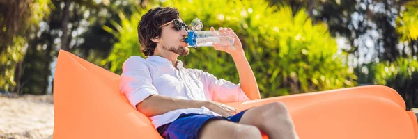 BANNER, LONG FORMAT Summer lifestyle portrait of men sitting on the orange inflatable sofa drinking water on the beach of tropical island. Relaxing and enjoying life on air bed — Stock Photo, Image