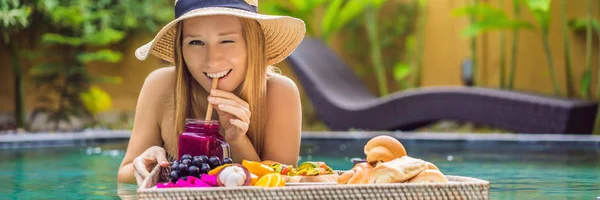 BANNER, LONG FORMAT Breakfast tray in swimming pool, floating breakfast in luxury hotel. Girl relaxing in the pool drinking smoothies and eating fruit plate, smoothie bowl by the hotel pool. Exotic