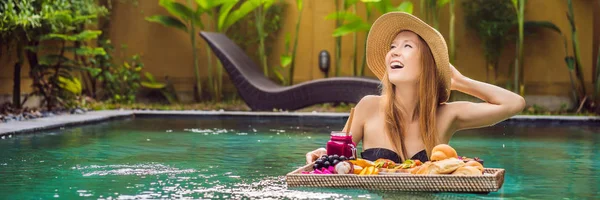 BANNER, LONG FORMAT Breakfast tray in swimming pool, floating breakfast in luxury hotel. Girl relaxing in the pool drinking smoothies and eating fruit plate, smoothie bowl by the hotel pool. Exotic