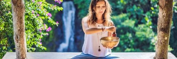 Mujer jugando en un cuenco tibetano mientras está sentada en una esterilla de yoga contra una cascada. Vintage tonificado. Hermosa chica con cuentas de mala meditación BANNER, FORMATO LARGO —  Fotos de Stock