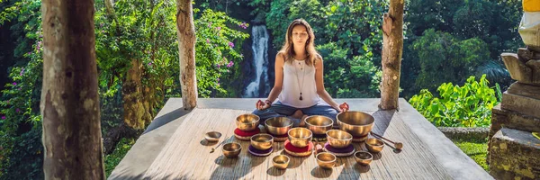 Mujer jugando en un cuenco tibetano mientras está sentada en una esterilla de yoga contra una cascada. Vintage tonificado. Hermosa chica con cuentas de mala meditación BANNER, FORMATO LARGO —  Fotos de Stock